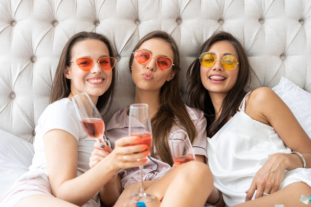Pajama party. Attractive young smiling women in pajamas drinking champagne while having a slumber party in the bedroom. - 写真・画像