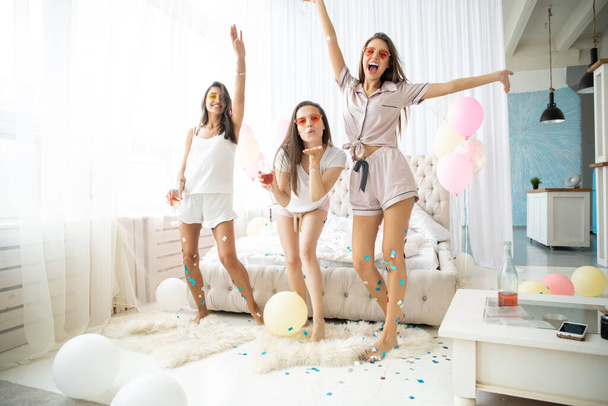 Three attractive young women in pajamas smiling and gesturing while jumping in bedroom with confetti flying everywhere - Photo, image