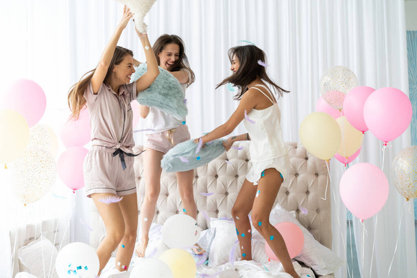 Three attractive young women in pajamas smiling and gesturing while jumping in bedroom with confetti flying everywhere - Φωτογραφία, εικόνα