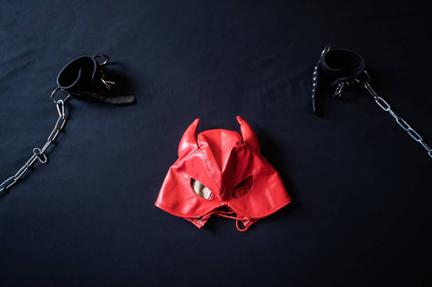 BDSM equipment. Top view of a red leather devil mask and black handcuffs with chains on a gray sheet. Sex toys. Dominance. No people. - Foto, afbeelding