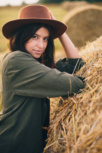 Stylish girl posing at hay bale in summer field in sunset. Portrait of young sensual woman in hat at haystack, atmospheric tranquil moment. Countryside slow life - Fotoğraf, Görsel