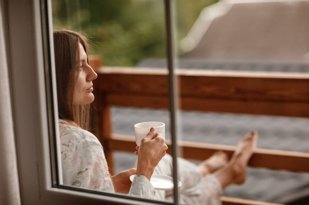 Young female standing after taking a shower in the morning on balcony of the hotel. holding a cup of coffee or tea in her hands. Looking outside nature forest and Mountain. - Foto, Imagen