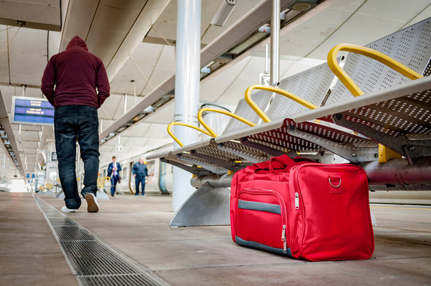 Terorismus a koncepce veřejné bezpečnosti s bezobslužnou taškou ponechanou pod židlí na nástupišti na vlakovém nádraží nebo na letišti a mužem v kapuci, který odchází od podezřelé věci (možná terorista) - Fotografie, Obrázek