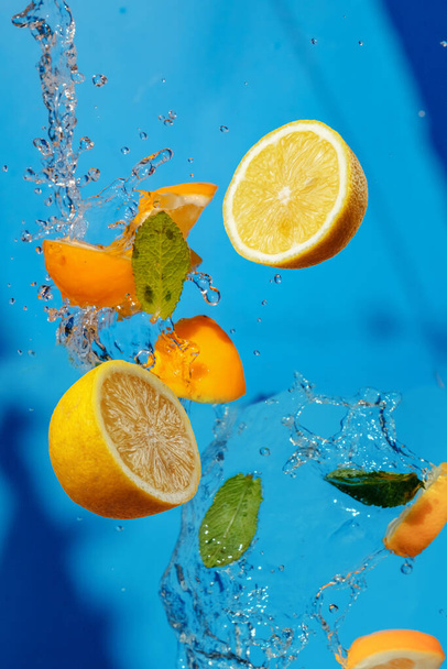 Падение свежих ломтиков лимона и зеленых листьев мяты с пресной водой в воздухе. Концепция летающих фруктов на голубом фоне  - Фото, изображение