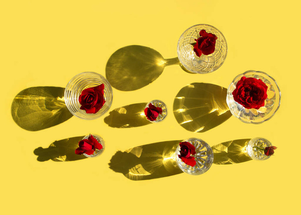 Γυαλιά με καθαρό νερό και κόκκινα τριαντάφυλλα στέκονται σε κίτρινο φόντο, πάνω όψη. Όμορφη δημιουργική νεκρή φύση πλημμυρίζει με φως με σκληρές σκιές και λάμψη από το γυαλί. Έννοια κόμματος. - Φωτογραφία, εικόνα