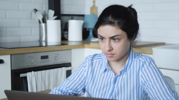 Μια σοβαρή νεαρή Ινδή με μπλε ριγέ πουκάμισο δουλεύει σε έναν υπολογιστή στο σπίτι και κάθεται σε μια φωτεινή κουζίνα. - Πλάνα, βίντεο
