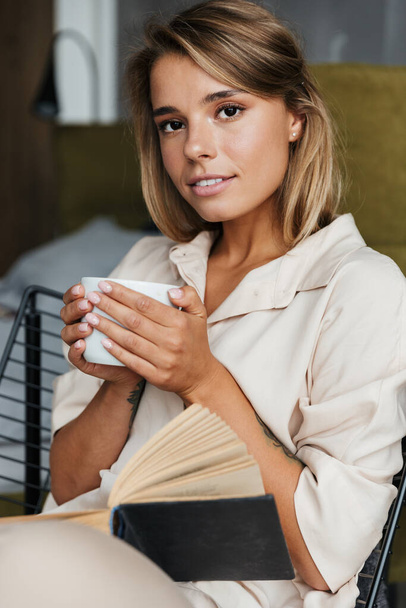 Image of young attractive nice woman drinking tea and reading book while sitting on armchair at living room - Photo, image