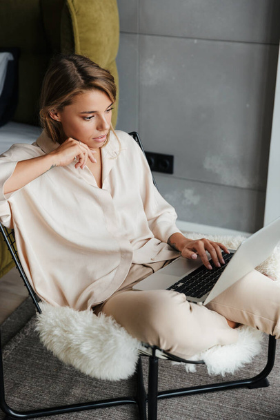 Image d'une belle femme blonde concentrée en pyjama élégant travaillant avec un ordinateur portable tout en étant assis sur un fauteuil au salon - Photo, image