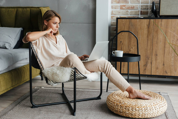 Image of focused blonde nice woman in stylish pajamas working with laptop while sitting on armchair at living room - Zdjęcie, obraz