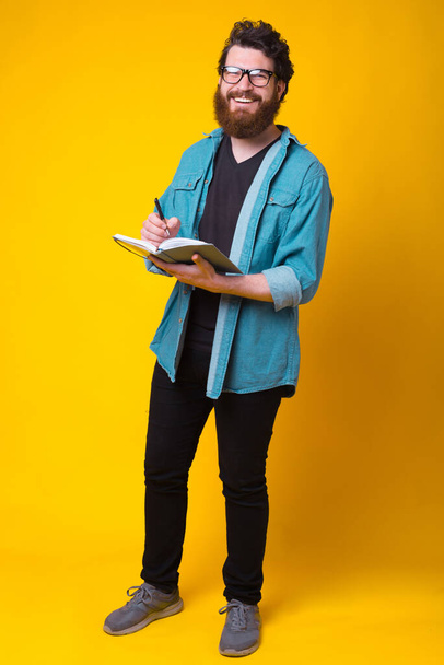 Felice barbuto hipster uomo sta sorridendo alla fotocamera mentre scrive nel suo diario su sfondo giallo. - Foto, immagini