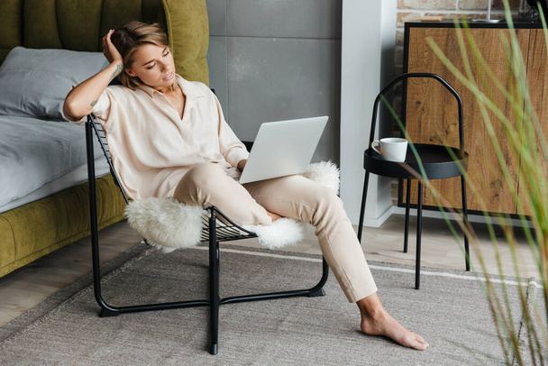 Image of focused blonde nice woman in stylish pajamas working with laptop while sitting on armchair at living room - Foto, Bild