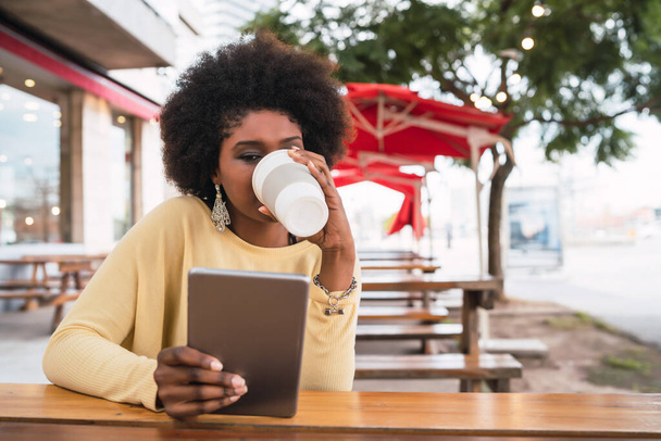 Portrait of young afro american latin woman using her digital tablet while sitting at the coffee shop. Technology concept. - 写真・画像