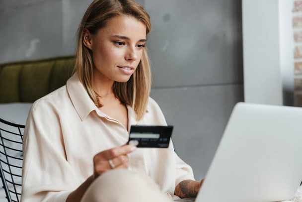 Image of focused nice woman in pajamas holding credit card and using laptop while sitting on armchair at living room - Photo, image
