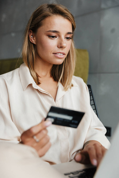 Image of focused nice woman in pajamas holding credit card and using laptop while sitting on armchair at living room - Fotoğraf, Görsel