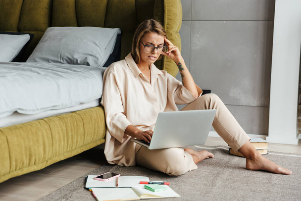 Image of perplexed nice woman in pajamas working with laptop and calendar while sitting on floor at bedroom - Foto, Bild