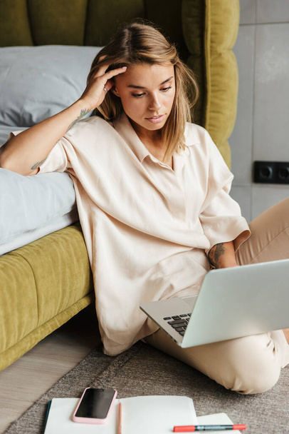 Image of serious nice woman in pajamas working with laptop and calendar while sitting on floor at bedroom - Photo, image