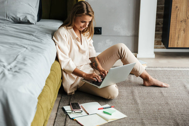 Image of serious nice woman in pajamas working with laptop and calendar while sitting on floor at bedroom - Foto, immagini