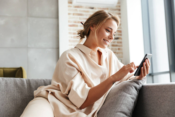 Image of happy nice woman in pajamas smiling and using cellphone while sitting on sofa at living room - Φωτογραφία, εικόνα