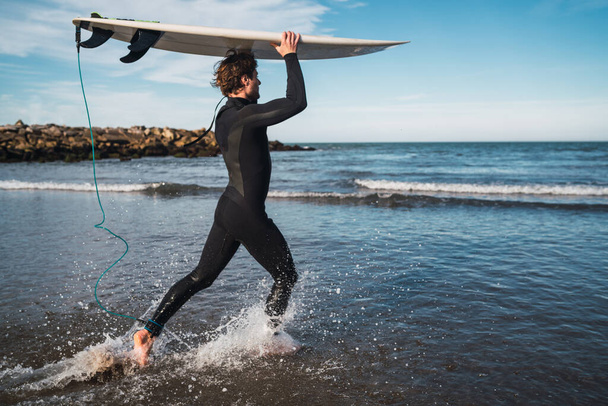 Young surfer entering into the water with his surfboard in a black surfing suit. Sport and water sport concept. - Foto, immagini