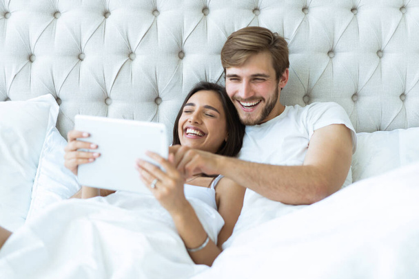 Happy young couple lying on the bed in a bedroom and using digital tablet. - Zdjęcie, obraz