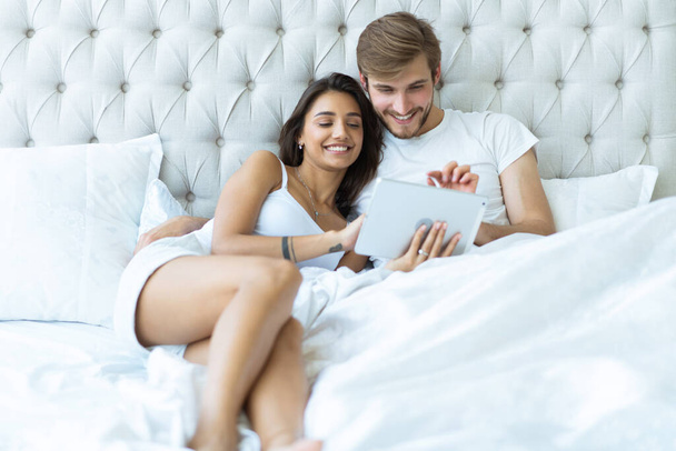 Happy young couple lying on the bed in a bedroom and using digital tablet. - Foto, Imagen