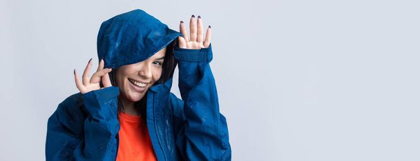 Muotokuva hymyilevä tyttö pukeutunut sininen sadetakki tippaa poseeraa huppu harmaalla taustalla studiossa. - Valokuva, kuva