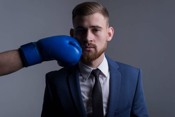 close-up portrait of a young bearded guy in a business suit, businessman, side view the hand of an opponent in a boxing glove strikes. The moment of punching in the face. Financial hit concept. competition. On a gray background. Business concept. Spo - Foto, Imagem
