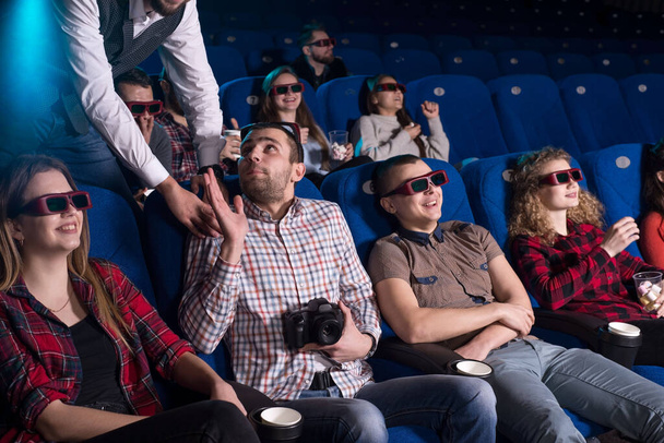 The cinema guard caught a young guy behind an illegal video while showing the film. Shooting pirates in the cinema. Against the background, people of different sexes are sitting, watching a movie in a movie theater, sitting in armchairs, eating popco - Fotografie, Obrázek