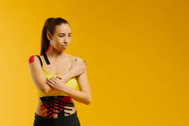 Young caucasian girl with kinesiology tape on her belly and leg, on yellow background. Sport and rehabilitation, kinesiotherapy treatment. - Foto, Bild