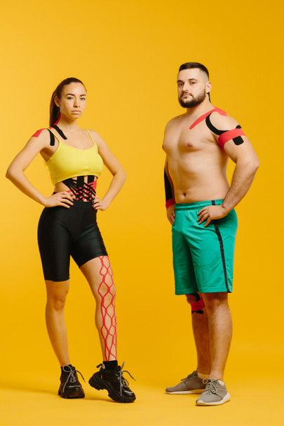 Profesionální sportovci, muž a žena s kineziologickou páskou na těle, pózující na žlutém pozadí. Sport a rehabilitace, kinezioterapeutická léčba - Fotografie, Obrázek