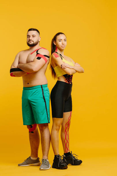 Professional athletes, man and woman with kinesiological tape on the body, posing on a yellow background. Sports and rehabilitation, kinesiotherapy treatment - Фото, изображение