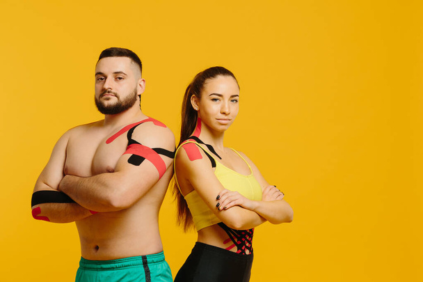 Profesionální sportovci, muž a žena s kineziologickou páskou na těle, pózující na žlutém pozadí. Sport a rehabilitace, kinezioterapeutická léčba - Fotografie, Obrázek