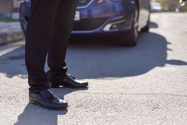 Männliche Beine in schwarzen Lederschuhen und schwarzer Hose, vor dem Hintergrund der Silhouette eines Autos in Unschärfe. - Foto, Bild