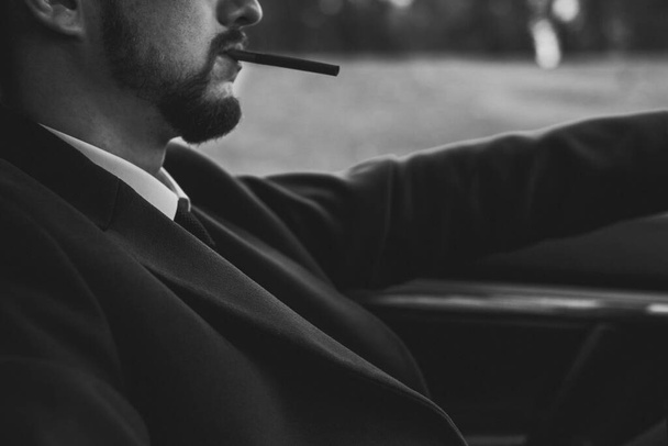 foto in bianco e nero, ritratto di un ragazzo di trent'anni, primo piano, che fuma una sigaretta mentre guida un'auto. Uomo d'affari con la barba in giacca e cravatta che fuma una sigaretta brutalmente. In elaborazione creativa. Un uomo barbuto fuma una cigare nera - Foto, immagini