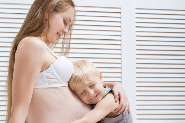 Little boy squint and hugging belly of pregnant mother. Young pregnant woman and son. - Photo, Image