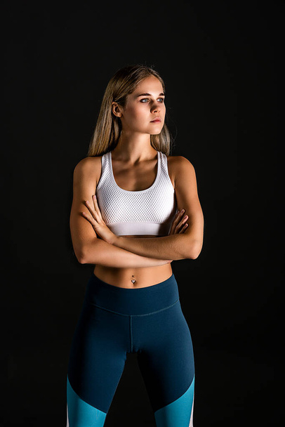 Halblanges Porträt eines blonden Mädchens in Sportuniform zum Fitnesstraining auf schwarzem Hintergrund. Arme vor dir auf Brusthöhe gefaltet - Foto, Bild