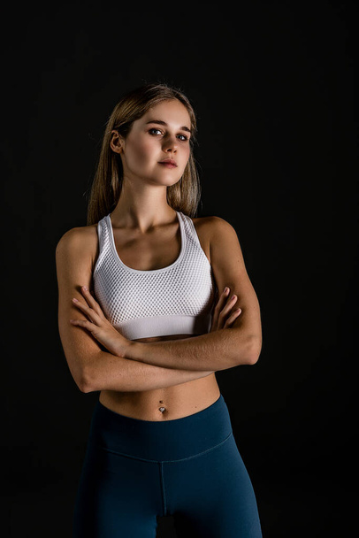Half-length portrait of a blonde girl in a sports uniform for fitness training on a black background. Arms folded at chest level in front of you - 写真・画像