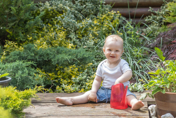 Smiling little baby boy is sitting in the garden holding  garden watering can and looking at the camera - Photo, image