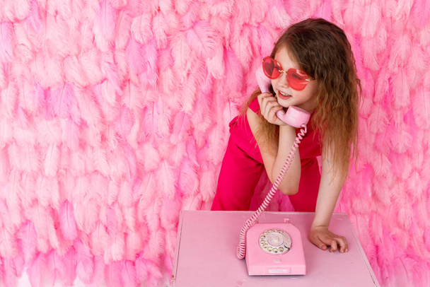 A little girl is sitting in stylish pink glasses on a pink background of their feathers, smiling a beautiful smile. A beautiful little girl is holding an old pink phone. Space for text - Fotografie, Obrázek