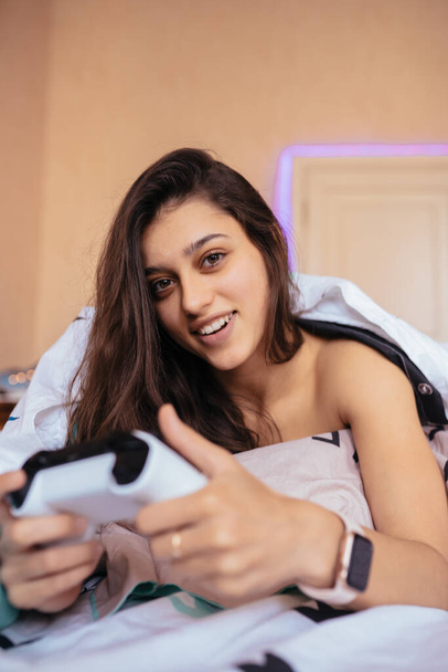 Funny girl lying in bed and playing video game, holding controller - Photo, image