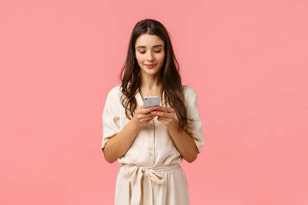 Ρομαντικό χαριτωμένο, όμορφη νεαρή γυναίκα στο φόρεμα, κουβεντιάζοντας με το φίλο, χαμογελώντας, κρατώντας smartphone και χτυπώντας οθόνη για να παραγγείλετε online, ψώνια χρησιμοποιώντας την εφαρμογή, στέκεται ροζ φόντο - Φωτογραφία, εικόνα