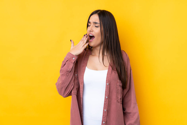 Young brunette woman over isolated yellow background yawning and covering wide open mouth with hand - Foto, imagen