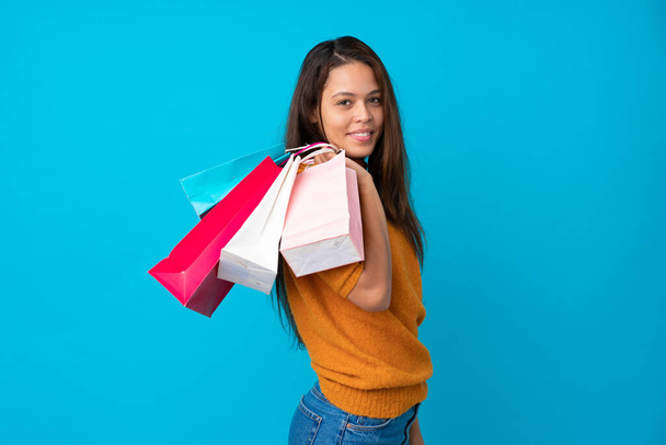 Молодая бразильская девушка на изолированном синем фоне с пакетами покупок и улыбкой
 - Фото, изображение
