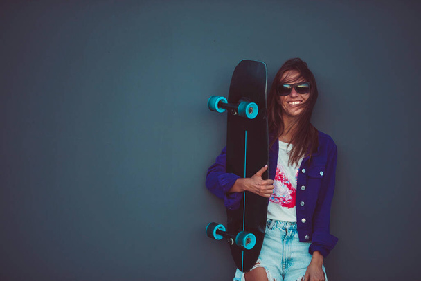 Young happy girl in sunglasses smiling with skateboard in front of grey wall. - 写真・画像