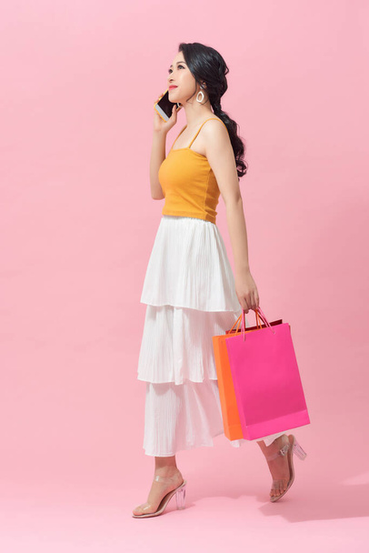 Πλήρες μήκος μιας όμορφης νεαρής γυναίκας που φοράει πολύχρωμα ρούχα και στέκεται απομονωμένη σε ροζ φόντο, κουβαλώντας τσάντες για ψώνια, κρατώντας κινητό τηλέφωνο - Φωτογραφία, εικόνα