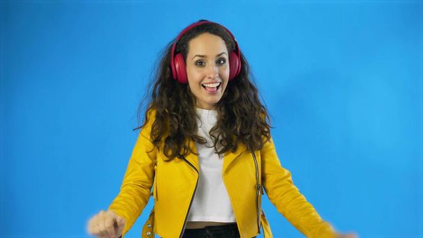 Young woman in yellow jacket dancing while listening music on headphones in Studio with blue Background. - Foto, afbeelding
