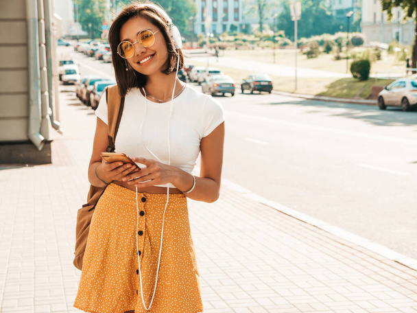 Fashion portrait of young stylish hipster woman walking in the street.Girl wearing cute trendy outfit.Smiling model enjoy her weekends, travel with backpack. Female listening to music via headphones - Photo, Image