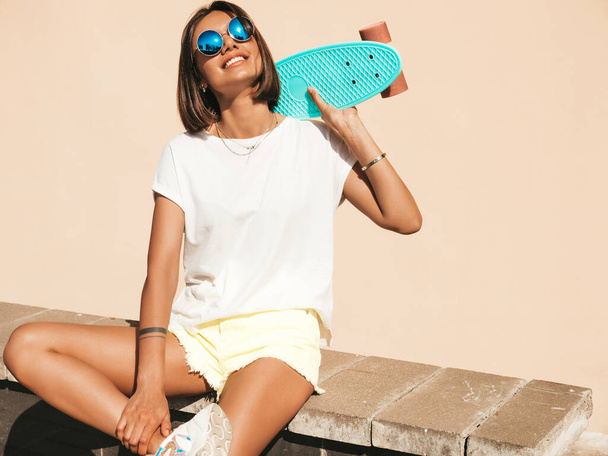 Young beautiful sexy smiling hipster woman in sunglasses.Trendy girl in summer T-shirt and shorts.Positive female with blue penny skateboard posing on the street background - Photo, Image