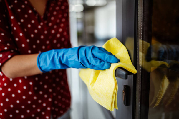 Νεαρή γυναίκα καθαρίζει το χερούλι της πόρτας με ένα κουρέλι που φοράει μπλε λαστιχένια γάντια. Απολύμανση γραφείου κατά την πανδημική καραντίνα. Έννοια της υγειονομικής περίθαλψης και του χώρου εργασίας - Φωτογραφία, εικόνα
