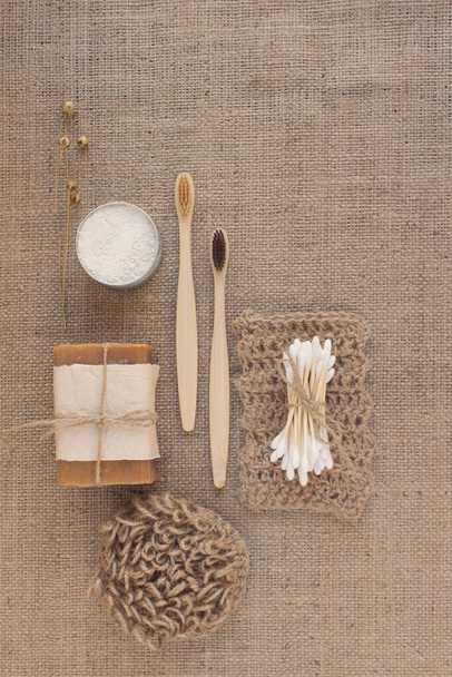 Vertical composition with natural bathroom tools, sustainable lifestyle concept. Wooden toothbrushes, bamboo swabs, organic soap, toothpowder, jute washcloth on fabric background with copy space - 写真・画像
